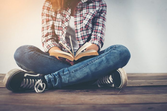 Young hipster woman holding open book sitting on wooden floor.